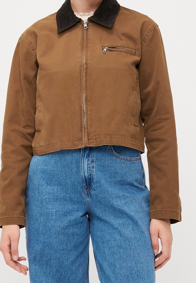 Chaqueta corta marrón con cuello de pana negro y bolsillo con cremallera, llevada sobre vaqueros azules de talle alto.