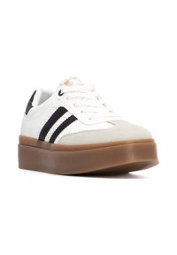 Zapatilla blanca con rayas negras, puntera de ante texturizado y suela de goma marrón gruesa. Presenta detalles texturizados y cordones planos.