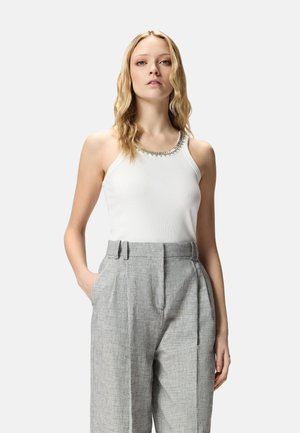 Donna che indossa un top bianco senza maniche con scollatura decorata con perline e pantaloni grigi su misura a vita alta, in piedi con una mano in tasca.
