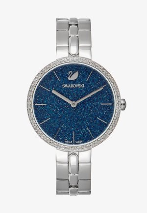 Reloj Swarovski con una caja metálica redonda de color plata, esfera azul con purpurina, marcadores de hora plateados y un bisel con cristales. Correa de acero inoxidable.