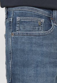 Jeans en denim bleu foncé avec un design à cinq poches, des coutures visibles et une petite étiquette en métal sur la poche arrière.
