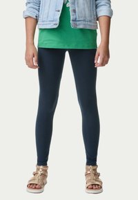 Leggings blu marina realizzati in tessuto elasticizzato, abbinati a una maglietta verde e a una giacca di jeans chiara. Le scarpe presentano cinturini dorati e suole bianche.