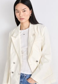 Dobbeltradet jakke i cremefarve, lavet af letvægtsstof, med kontrasterende syninger, to forlommer og sølvfarvede hardware-detaljer.