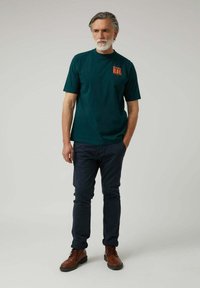 Camiseta de algodón verde oscuro con el texto "DREAM BIG" en naranja en el lado izquierdo del pecho, combinada con pantalones azul marino y botas marrones con cordones.