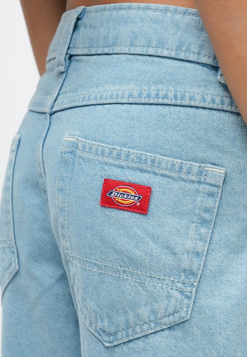 Lichtblauwe spijkerbroek met een achterzak waarop aan de rechterkant een rood-gele Dickies merklogo-patch is bevestigd.
