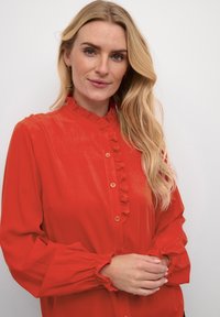 Red blouse made of soft fabric, featuring a ruffled collar, button closure, and gathered sleeves. Smooth texture with subtle sheen.