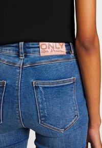 Gros plan sur un jean en denim bleu avec un patch en cuir marron étiqueté "ONLY Blue Denim", porté avec un haut noir, montrant les poches arrière et le bras latéral.