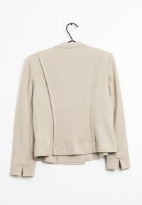 Béžové na míru šité sako s hladkou texturou, dvěma zadními panely a rozparkem na rukávech. Visí na černém háčku na bílém pozadí.