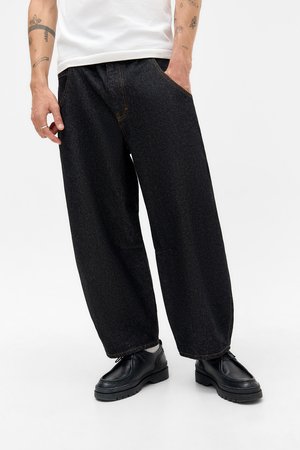 Persoon in losse zwarte spijkerbroek, zwarte leren schoenen en een wit shirt, staand met één hand in de zak tegen een witte achtergrond.