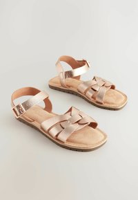Sandalias de oro rosa con tiras trenzadas, hebilla ajustable en el tobillo, plantilla de tono marrón suave y suela de goma texturizada.