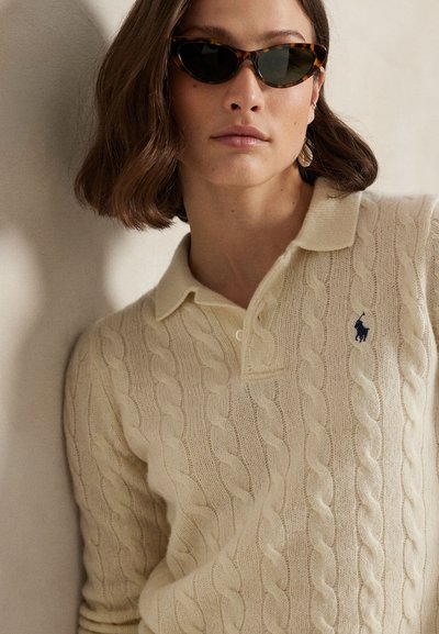 Vrouw die een schildpad-kattenoog-zonnebril draagt en een crème kabelgebreide polo trui met geborduurd logo, tegen een beige achtergrond.