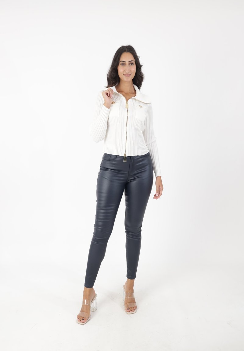 Chaqueta blanca de punto con cremallera y cuello, con dos bolsillos en la parte frontal; combinada con leggings de imitación de piel negros y sandalias de tacón bloque transparente.