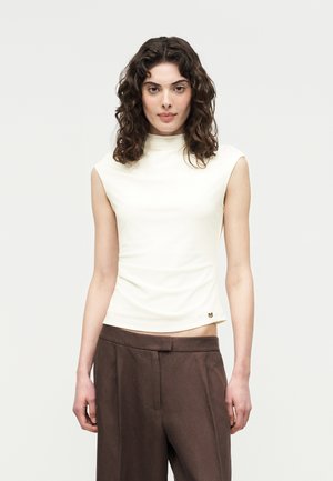 Μια γυναίκα με σγουρά σκουρόχρωμα μαλλιά φοράει ένα λευκό αμάνικο τοπ με ψεύτικο γιακά και ψηλόμεσα καφέ παντελόνια, στέκεται μπροστά σε ένα απλό ανοιχτόχρωμο φόντο.