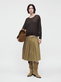 Femme portant un haut noir à manches longues, une jupe plissée couleur olive, des bottes marron décontractées, tenant un grand sac en daim marron sur un fond uni.