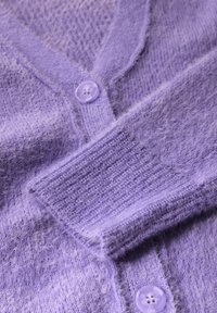 Lila kofta gjord av mjukt, strukturerat tyg med V-hals, ribbade manschetter och två lila knappar framtill.