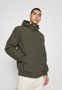 Maslinasto zeleni zip-up kaput s kapuljačom, sa elastičnim manžetama, dva bočna džepa i suptilnim logom sprijeda. Glatka, lagana tkanina.