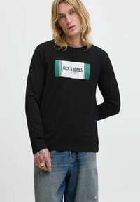 Eine blonde Person trägt ein schwarzes Langarmshirt mit dem Logo "JACK & JONES COLLECTIONS" und blaue Jeans, eine Hand hinter dem Rücken.