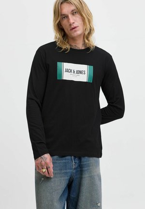 Blond osoba u crnoj majici dugih rukava s logom "JACK & JONES COLLECTIONS" i plavim trapericama, s jednom rukom iza leđa.