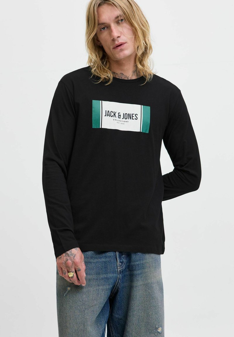 Eine blonde Person trägt ein schwarzes Langarmshirt mit dem Logo "JACK & JONES COLLECTIONS" und blaue Jeans, eine Hand hinter dem Rücken.