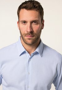 Camisa de botón azul claro con un pequeño patrón a cuadros, diseño con cuello y una tela texturizada. Presenta botones claros y una forma ajustada.