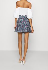 Haut blanc à épaules dénudées avec des manches bouffantes, associé à une jupe évasée navy à fleurs avec des motifs roses et verts, et des mules blanches à talons.