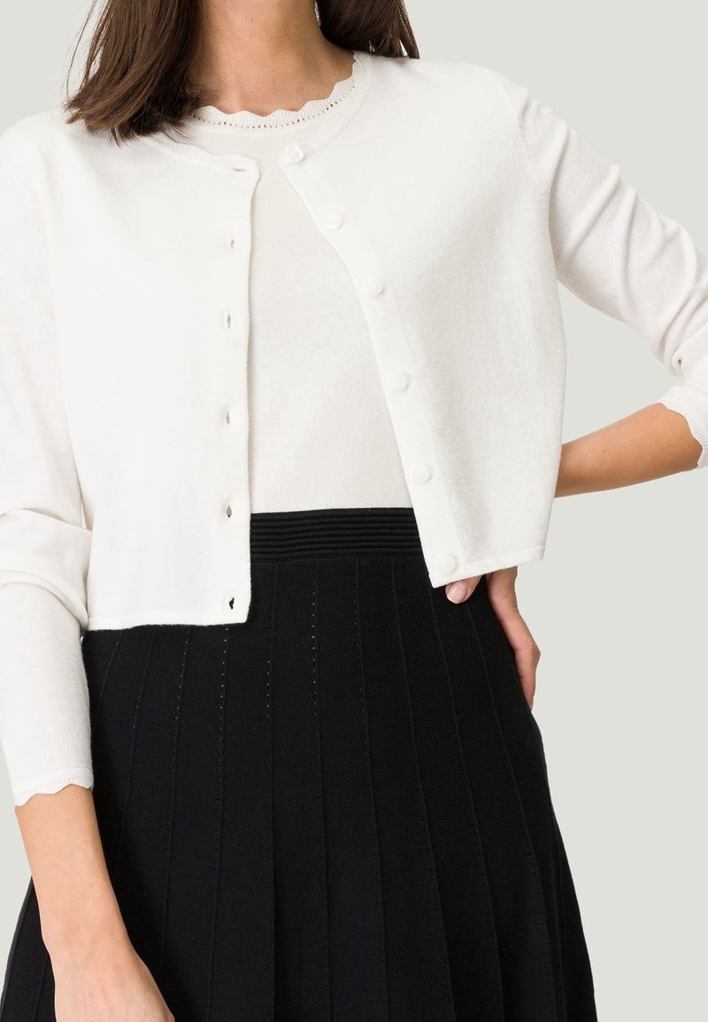 Femme portant un cardigan court blanc boutonné sur un top blanc aux bords festonnés, associé à une jupe noire plissée taille haute.