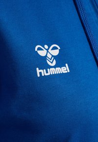 Blauwe stof met een gladde textuur toont een wit logo van een gestileerde bij boven de tekst "hummel" in vet, modern lettertype.