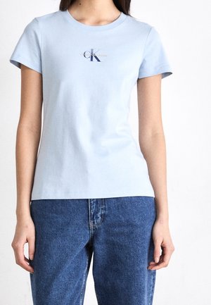 Vrouw die een lichtblauw Calvin Klein T-shirt met korte mouwen en blauwe spijkerbroek draagt, staand tegen een witte achtergrond.