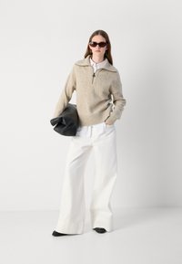 Suéter de malha bege com gola com zíper, combinado com uma camisa branca e calças largas brancas. Bolsa de mão estruturada preta e óculos de sol com tonalidades.