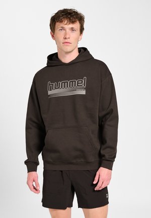 Młody mężczyzna w czarnej bluzie z kapturem "hummel" i czarnych szortach, stojący na jednolitym jasnoszarym tle.