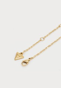 Gouden halsketting met een driehoekige hanger gegraveerd met "GUESS" en een kreeftsluiting. Beschikt over een delicate schakelkettinkje.