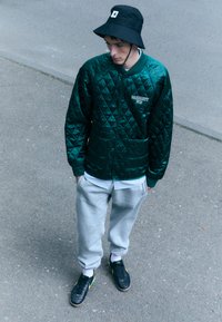 Quilted green satin jacket with pockets, worn over a light blue shirt, paired with gray sweatpants and black sneakers with yellow accents.