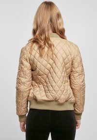 Quiltet beige bomberjakke med ribbestrikket kant og mansjetter, som har et diamantmønster og en glatt tekstur. Bakfra vises minimal detaljering.