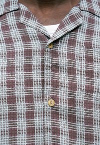 Uomo che indossa una camicia a quadri marrone e bianca con colletto aperto e una maglietta bianca visibile sotto.