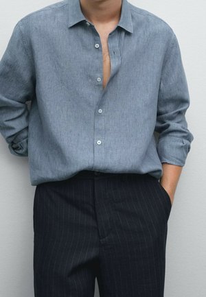 Homme portant une chemise bleu clair à manches longues avec des boutons partiellement déboutonnés et un pantalon sombre à fines rayures, les mains dans les poches.