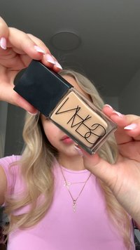 Flacon de fond de teint avec un corps en verre transparent et un bouchon noir mat. Liquide beige clair visible à l'intérieur. L'étiquette présente des lettres noires en gras.