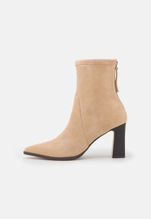 Beige ruskind ankelstøvle med spids tå, blokhæl og lynlås bagpå, vist i profil mod en ensfarvet baggrund.