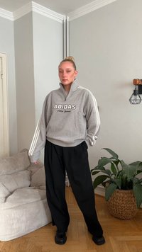 Sweatshirt de lã cinzenta com fecho de correr e riscas brancas, com o logótipo "ADIDAS". Combinada com calças largas pretas e sapatos pretos. Ambiente interior.