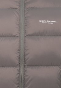 Sivá prešívaná bunda s výzipom, prešívanou textúrou a logom "ARMANI EXCHANGE" v bielej farbe. Hladká tkanina s matným povrchom.