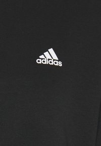 Logo Adidas blanc avec trois bandes et texte sur fond de tissu noir, centré dans le cadre.