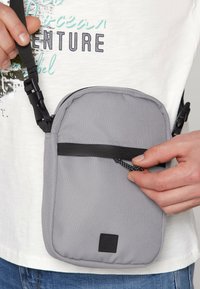 Graue Stoff-Umhängetasche mit einer Reißverschlusstasche, strukturiertem Oberflächen, schwarzem Tragegurt und einem kleinen Markenpatch auf der Vorderseite.