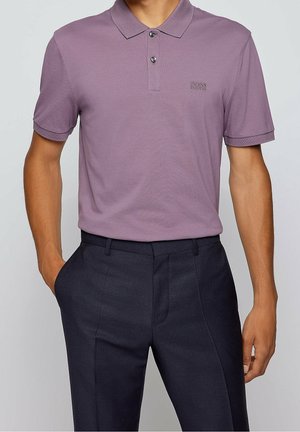 Homme portant un polo mauve à manches courtes avec des boutons et un pantalon foncé sur mesure, une main dans la poche, debout devant un fond uni.