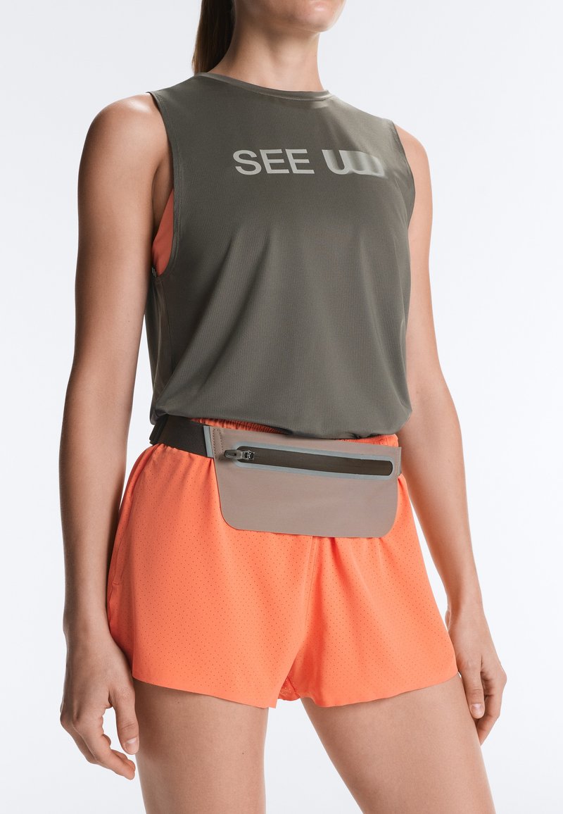 Femme portant un top sans manches vert olive avec le texte "SEE", un short orange vif et une pochette beige zippée à la taille sur fond blanc.
