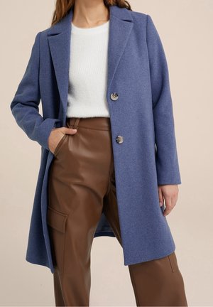 Kobieta w niebieskim płaszczu zapinanym na guziki, na białym swetrze i brązowych skórzanych spodniach cargo, z jedną ręką w kieszeni na beżowym tle.