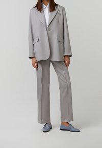 Helles graues Anzug mit einem leicht übergroßen Blazer, der einen einzelnen Knopf und zwei Fronttaschen hat, kombiniert mit maßgeschneiderten Hosen und blauen Slippern.
