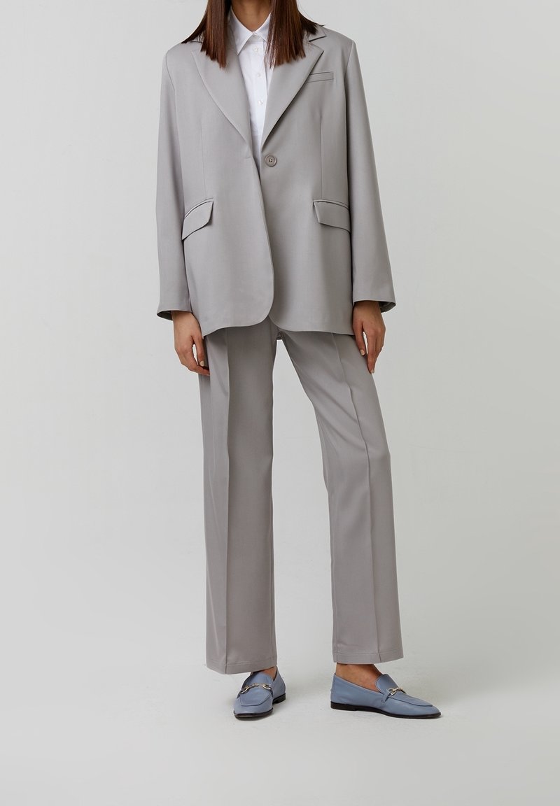 Helles graues Anzug mit einem leicht übergroßen Blazer, der einen einzelnen Knopf und zwei Fronttaschen hat, kombiniert mit maßgeschneiderten Hosen und blauen Slippern.