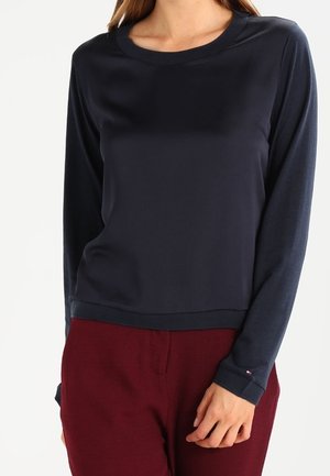 Vrouw draagt een marineblauwe top met lange mouwen en ronde hals en bordeauxrode broek, staand tegen een witte achtergrond.