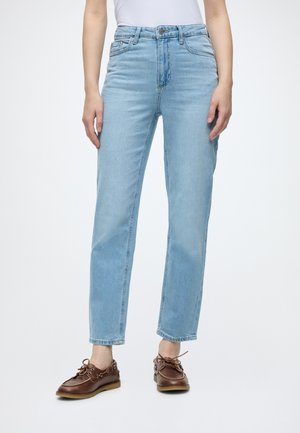 Femme portant un jean bleu clair droit, une chemise blanche et des chaussures bateau en cuir marron à lacets, debout sur fond blanc.