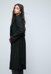 Giovane donna con lunghi capelli scuri indossa un lungo cappotto nero con una cintura, in piedi di lato contro uno sfondo grigio chiaro uniforme.