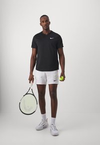 Zwarte Nike T-shirt met korte mouwen, witte shorts met zwart logo, houdt een tennisracket en een gele tennisbal vast. Witte sportschoenen en sokken.